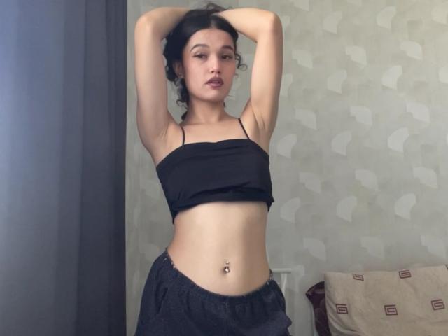 YoulandaCarlough with arms raised, wearing a black crop top and dark pants, showing a flat stomach with a navel piercing, looking at the camera.