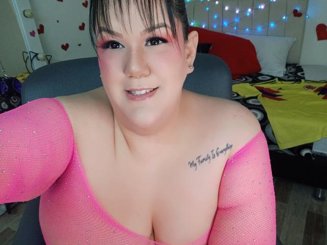 GiiannaMorrys with pink mesh top, large tits, tattoo on right shoulder, smiling, pink eye makeup, black hair, in chair, bedroom with string lights and heart decorations.