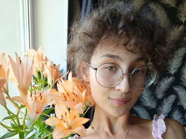 Alyce Erway, con cabello castaño rizado, gafas y una pequeña sonrisa, está de pie junto a un ramo de lirios naranjas. Sus senos son pequeños y lleva una cinta rosa..