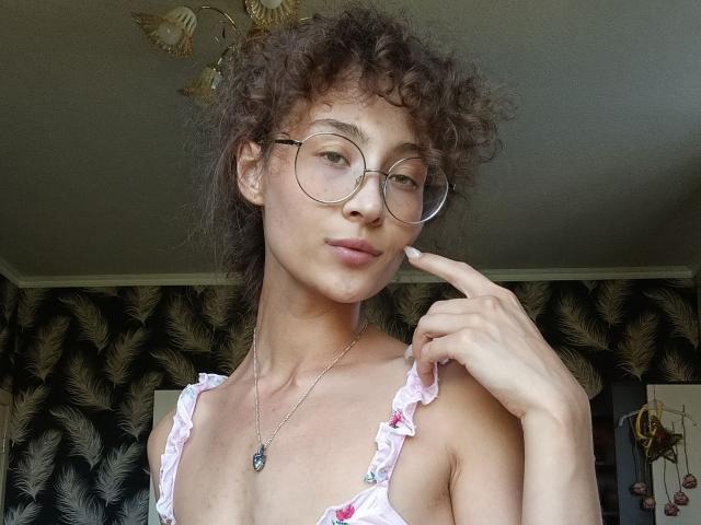 AlyceErway, con i capelli castano ricci, gli occhiali e una maglietta rosa con volant, indica le sue labbra leggermente dischiuse, mostrando un'espressione carina e sicura di sé, indossando una collana con un ciondolo a forma di cuore..