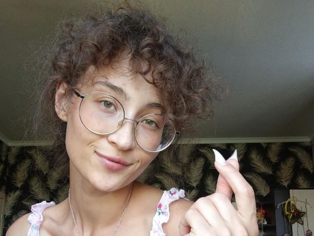 Curly-haired AlyceErway with glasses, pink floral top, showing white-painted nails, smiling slightly, light skin, small tits, slim build, holding up her hand in a cute gesture.