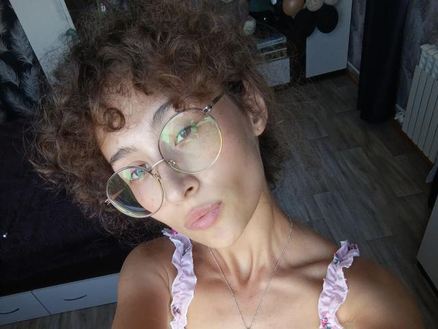 AlyceErway with curly brown hair, wearing round glasses and a pink frilled top, has a pouty expression and a necklace, showing her tits and a slim neck.
