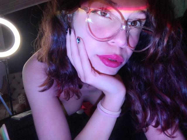 SophieBlake with red lipstick, pink glasses, and wavy brown hair, resting her chin on her hand, showing her tits, dark nails, and a ring on her finger.
