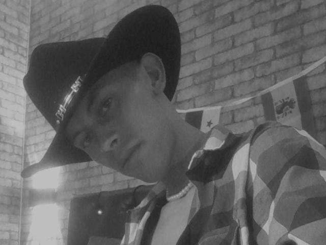 Fotografía en blanco y negro de AnderCooper con un sombrero de vaquero, vistiendo una camisa a cuadros, un collar de perlas y una expresión seria. AnderCooper tiene una complexión media y una nariz ligeramente puntiaguda..