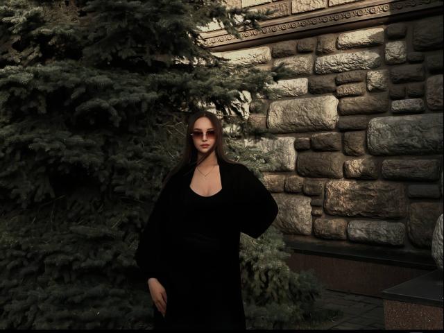 TawnyaKaczmarski stands with hands on hips, wearing a black dress, long sleeves, and a necklace, in front of a stone wall and large green pine tree.