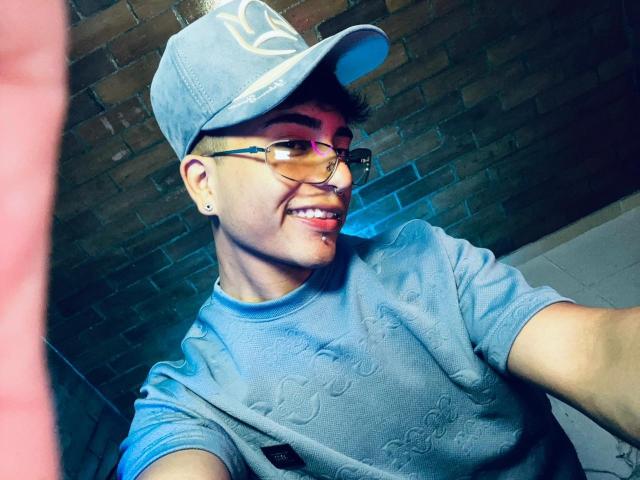 KermyBerty wearing a blue baseball cap, glasses, and a blue shirt with subtle patterns, smiling with a slight nose piercing, and visible arm in a selfie.