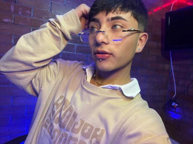 KermyBerty has short dark hair, glasses, and a nose ring. Wearing a beige sweater, white collared shirt, and a lip piercing, with a blue strip on his cheek.