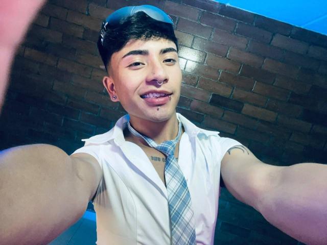 KermyBerty with short black hair, wearing a white sleeveless shirt, blue plaid tie, and glasses on head, smiling with tongue out, nose ring, and tattoos, taking a selfie.