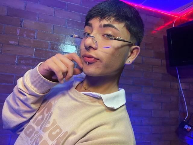 KermyBerty, wearing glasses and a beige polo shirt, sticks out tongue, with nose ring and facial piercing, in dim blue and red lighting, holding finger to chin.