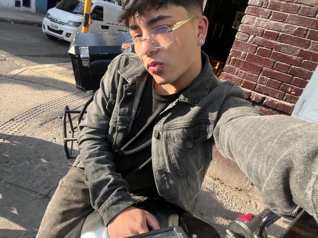 KermyBerty with short dark hair, wearing glasses and a grey denim jacket, pouting with lips slightly parted, sitting on a motorcycle, holding the handlebars.