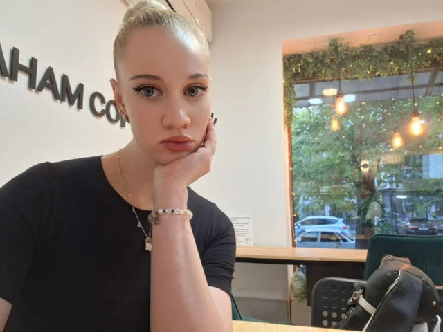 LindaxxCrow con chignon biondo, camicia nera, mano sul viso, che guarda la fotocamera, trucco leggero, collana, braccialetto, seduta in un caffè, sedia verde, borsa nera, luce soffusa, espressione seria..