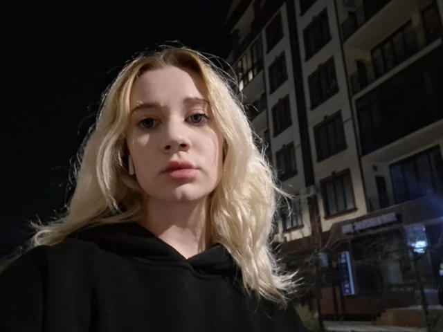 LindaxxCrow with blonde hair, wearing a black hoodie, has a serious expression, and is in a night-time urban setting with dimly lit buildings in the background.