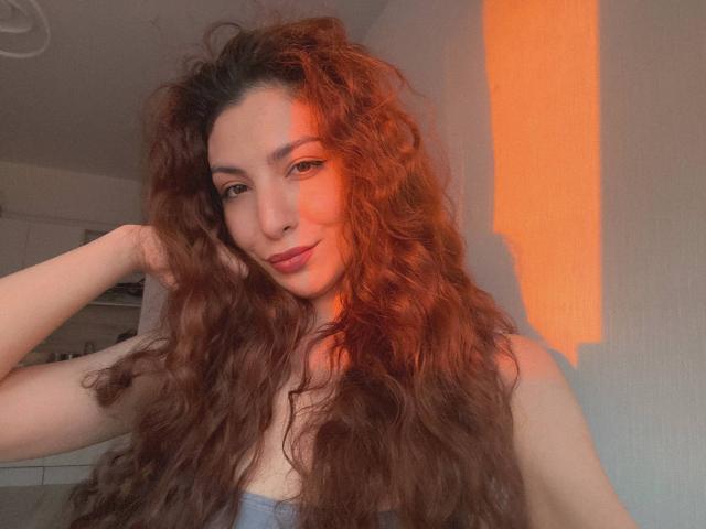 NensyxGenry, con cabello castaño largo y rizado, piel clara y una ligera sonrisa, vestida con una blusa gris, con la mano en el cabello, luz solar naranja en el lado derecho de su rostro..