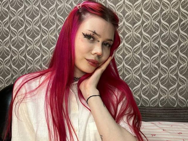ToriHananocha met felroze haar, gekleed in een witte blouse, een zwarte armband en een choker, met haar wang op haar hand rustend, waarbij een neusring en subtiele make-up zichtbaar zijn..