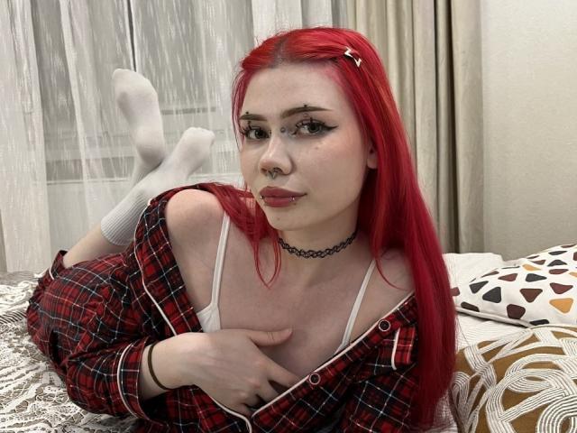 ToriHananocha, avec des cheveux roux, un anneau au nez et un collier noir, allongée sur un lit, portant une chemise à carreaux rouges, un soutien-gorge blanc et des chaussettes blanches, une main sur la poitrine, regardant l'appareil photo..