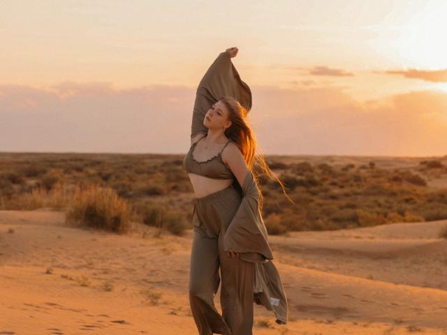 StellaLane, con lunghi capelli rossi, indossa un reggiseno e dei pantaloni verdi, alza un braccio, la luce del sole la illumina, è in piedi in un deserto sabbioso, appare felice e libera..