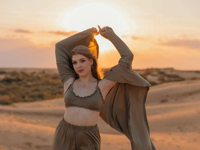 StellaLane, con piccoli seni, indossa un reggiseno verde e pantaloni larghi, alza le braccia togliendosi una giacca verde, tramonto in un deserto sabbioso, cielo arancione, lunghi capelli castani, espressione calma..