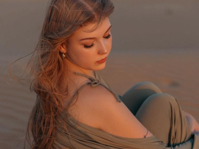 StellaLane, con lunghi capelli castani, guarda in basso, e i suoi seni sono leggermente scoperti sotto una maglia verde a spalle scoperte. È seduta su una spiaggia sabbiosa, indossa pantaloni verdi e orecchini dorati..