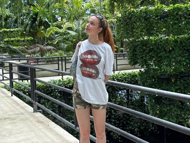 Joette Christianson, con cabello rojo recogido en una coleta, vestida con una camiseta blanca con un estampado de un labio rojo, pantalones cortos de camuflaje y gafas de sol en la cabeza, de pie junto a una barandilla negra..