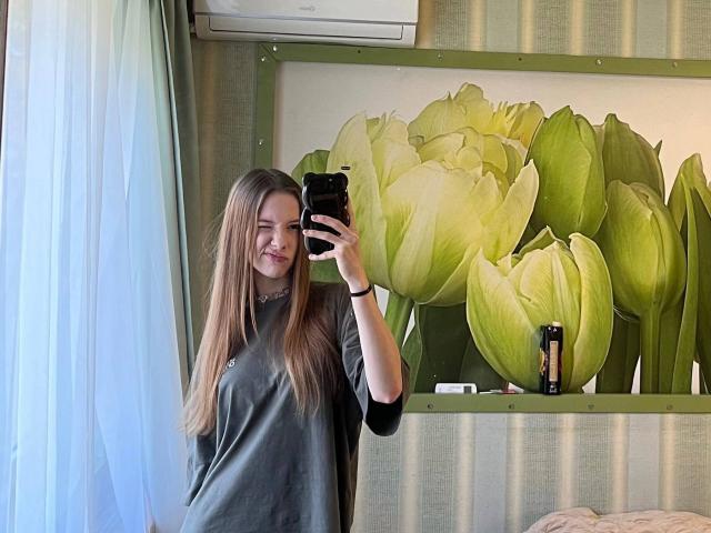 Joette Christianson, com cabelos castanhos longos, sorrindo, vestindo uma camisa cinza, tirando uma selfie no espelho com um celular preto, em pé perto de uma janela, em um quarto com papel de parede listrado de verde..