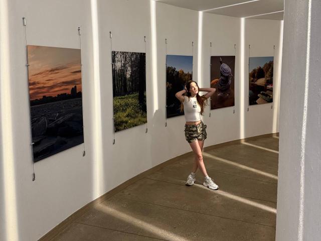 Joette Christianson staat in een kunstgalerie, gekleed in een wit tanktop, camouflage shorts, witte sneakers, en ze heeft haar lange bruine haar om haar handen, met vijf ingelijste foto's aan de muren..