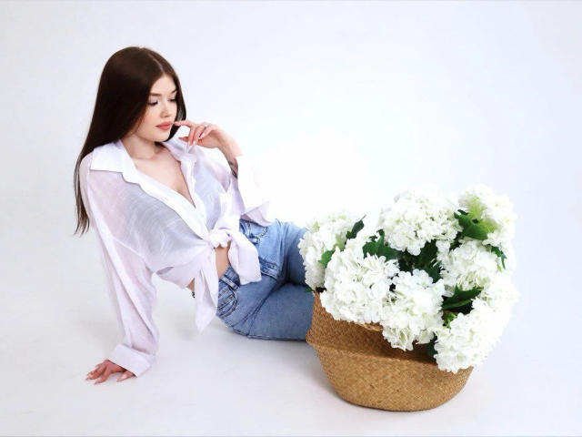 TrudieProse, mit langem, braunem Haar, trägt ein weißes, hochgekrempeltes Hemd und blaue Jeans und sitzt neben einem Korbkorb mit weißen Chrysanthemen..
