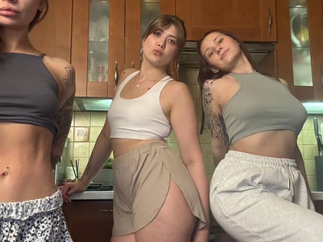 Three HotCaramelsTrio standing in a kitchen, wearing crop tops and high-waisted shorts. Their toned bellies and tattoos are visible. One has gray shorts, another has beige shorts.