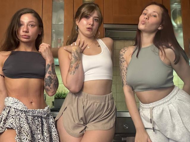 Three HotCaramelsTrio women standing in a kitchen, all with tattoos, wearing crop tops and shorts, showing toned midriffs, and making playful faces. Boobs, ass, and tattoos visible.
