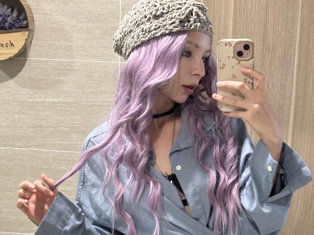 ChloeMoreaux with lilac wavy hair, wearing a knitted beanie and blue shirt, holding her hair and taking a mirror selfie, showing her black choker and phone with floral case.