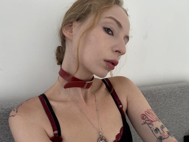 ChloeMoreaux with light brown hair in a messy bun, red collar, black and red lace bra, lip piercing, tattoos on her arm, sitting on a gray couch, looking side.
