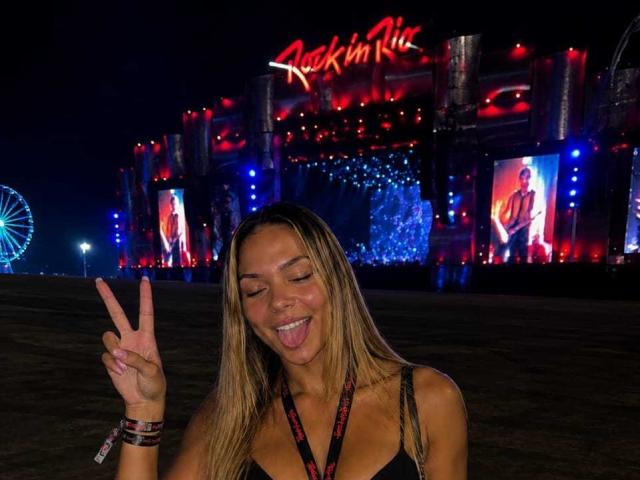 UrsulaX with long blonde hair, tongue out, and eyes closed, making a peace sign, wearing a black top, and a red-and-black necklace, in front of a neon-lit building.