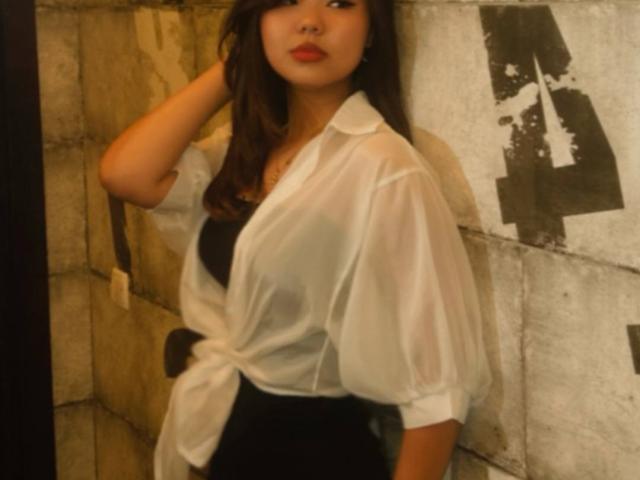 Ella Badgero, com cabelos castanhos longos, batom vermelho, vestindo uma blusa branca transparente amarrada na cintura, uma blusa preta por baixo, em pé contra uma parede, olhando para o lado, com a mão no cabelo..