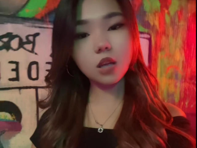 EllaBadgero, avec de longs cheveux bruns, la bouche légèrement ouverte, une lumière rouge sur le visage, portant une chemise noire et un collier, regardant vers l'avant. Ses seins sont discrets mais visibles..