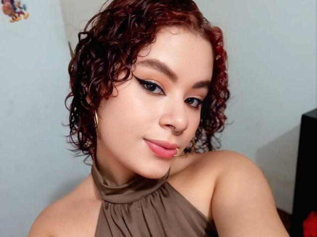 VeronicaMarie, com cabelos ruivo-acastanhados cacheados, blusa de alça marrom, brincos grandes, piercing no nariz e delineador, sorrindo para a câmera. Seus seios são pequenos e suas nádegas não estão visíveis..