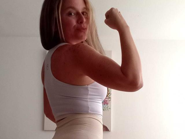 PamelaColin, flexing her right arm, has a cute smile, and is wearing a white sports bra with beige high-waisted pants, showing her toned, muscular ass.