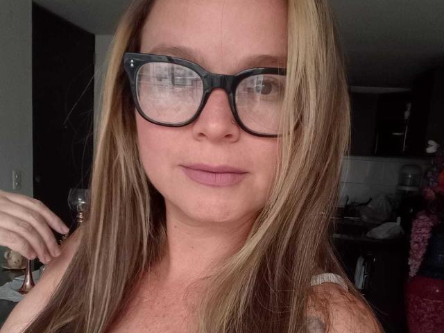 PamelaColin with long blonde hair, wearing black glasses, and pink lipstick, has a neutral expression, and is holding a wine glass, showing her tits slightly.