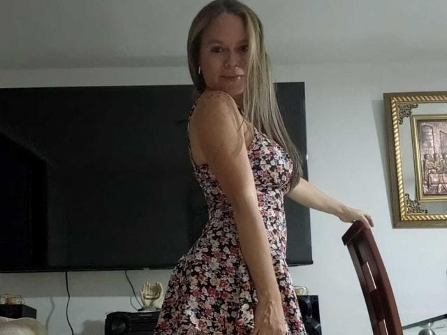 PamelaColin with long brown hair, wearing a floral dress, standing sideways, showing her ass, holding a chair, smiling, in a room with a TV and framed picture.