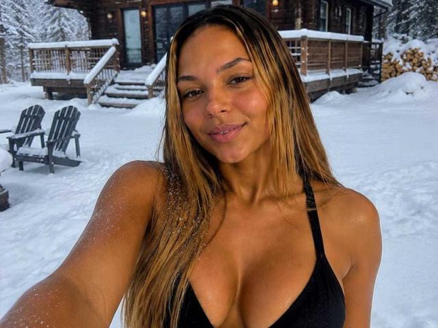 UrsulaX with long brown hair, tan skin, and big tits in a black bikini, standing in snow, smiling at the camera, with sand on her arm.
