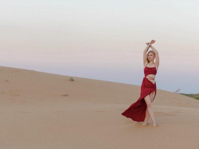NicoleBrian, con le braccia alzate, indossa un abito rosso con tagli e spacchi, in piedi in un deserto sabbioso, con le gambe e i piedi scoperti..