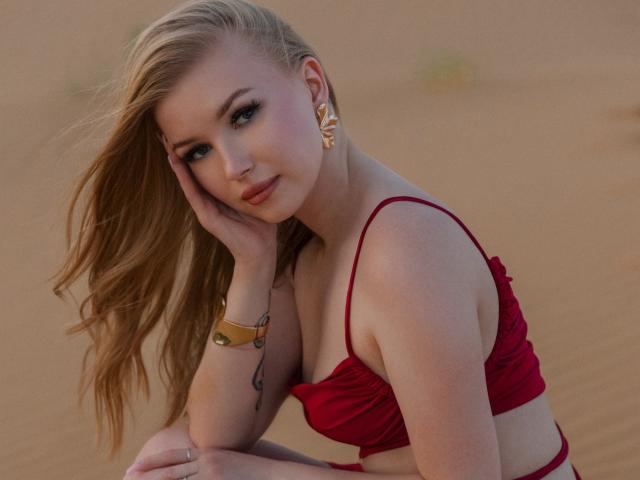 NicoleBrian, con i capelli biondi, un top da bikini rosso, orecchini dorati e un braccialetto, appoggia la guancia sulla mano, sorridendo, mostrando il seno, in un deserto..