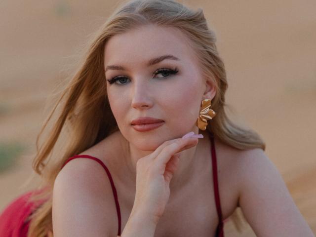 Nicole tem cabelo loiro, olhos verdes e está usando brincos de folha dourada. Ela está vestindo uma blusa vermelha, com a mão embaixo do queixo, e parece sedutora..