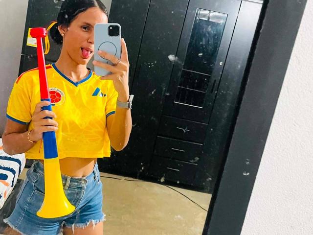 LaBandy, avec ses cheveux foncés attachés en couettes, la langue tirée, portant un haut court jaune et un short bleu, tenant un jouet jaune et rouge, prenant un selfie devant un miroir..