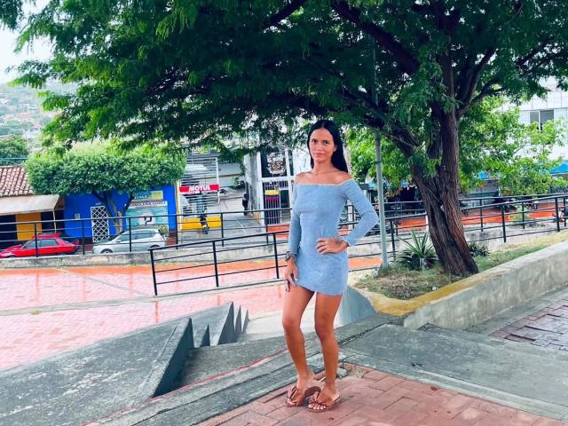 LaBandy se tient debout, vêtue d'une robe bleue à une épaule, qui révèle sa poitrine et ses fesses, et porte des sandales marron. Elle se trouve sous un arbre, sur une rue pavée de briques, et sourit, une main sur sa hanche..