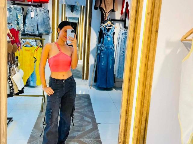 LaBandy vestindo uma cropped vermelha, mostrando os seios, e calças cargo pretas, tirando uma selfie no espelho em uma loja de roupas, com jeans e vestidos ao fundo..