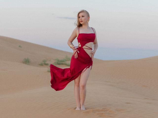 NicoleBrian è in piedi nel deserto, a piedi nudi, indossando un abito rosso con tagli che mostrano il seno e una spaccatura che rivela la vulva. Il suo abito svolazza nel vento..
