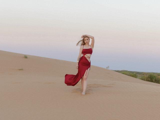 NicoleBrian, barfuß, tanzend in einer Wüste, trägt ein rotes, fließendes, trägerloses Kleid mit einem hohen Schlitz, der ihre Oberschenkel freilegt, und hält ihr Haar, mit einem friedlichen Ausdruck..