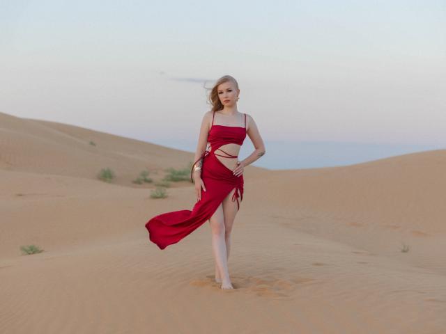 NicoleBrian in einem roten, fließenden Kleid mit Ausschnitten, barfuß in einer Wüste stehend, Wind weht durch ihr blondes Haar, Kleid wirbelt, blasse Haut, schlank, kleine Brüste, subtiles Lächeln..