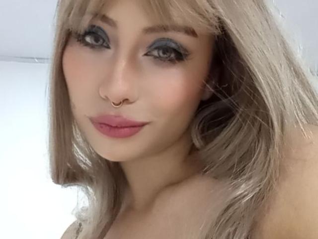 Ilaynna with blonde hair, gray eye makeup, pink lipstick, nose ring, and soft smile. Her tits are slightly visible, and she's looking directly at the camera.