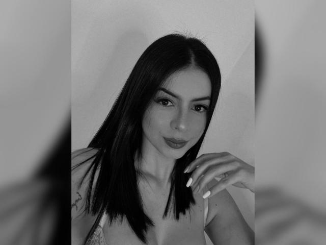 Foto em preto e branco de Isabella James com cabelo preto e liso, uma tatuagem discreta no ombro esquerdo, vestindo uma blusa branca, olhando para a câmera com um leve sorriso, com a mão direita levantada..