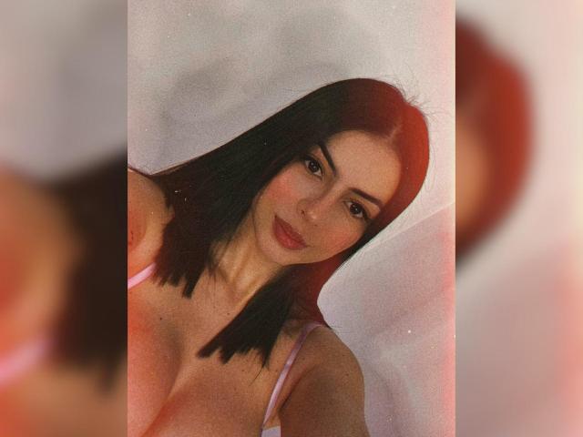 Isabella James mit schulterlangem, schwarzem Haar, rosafarbenen BH-Trägern und einem leichten Lächeln, die ihre Brüste zeigt, mit einem weichen, unscharfen, rot getönten Hintergrund. Ihre Haut ist hell und sie ist attraktiv..